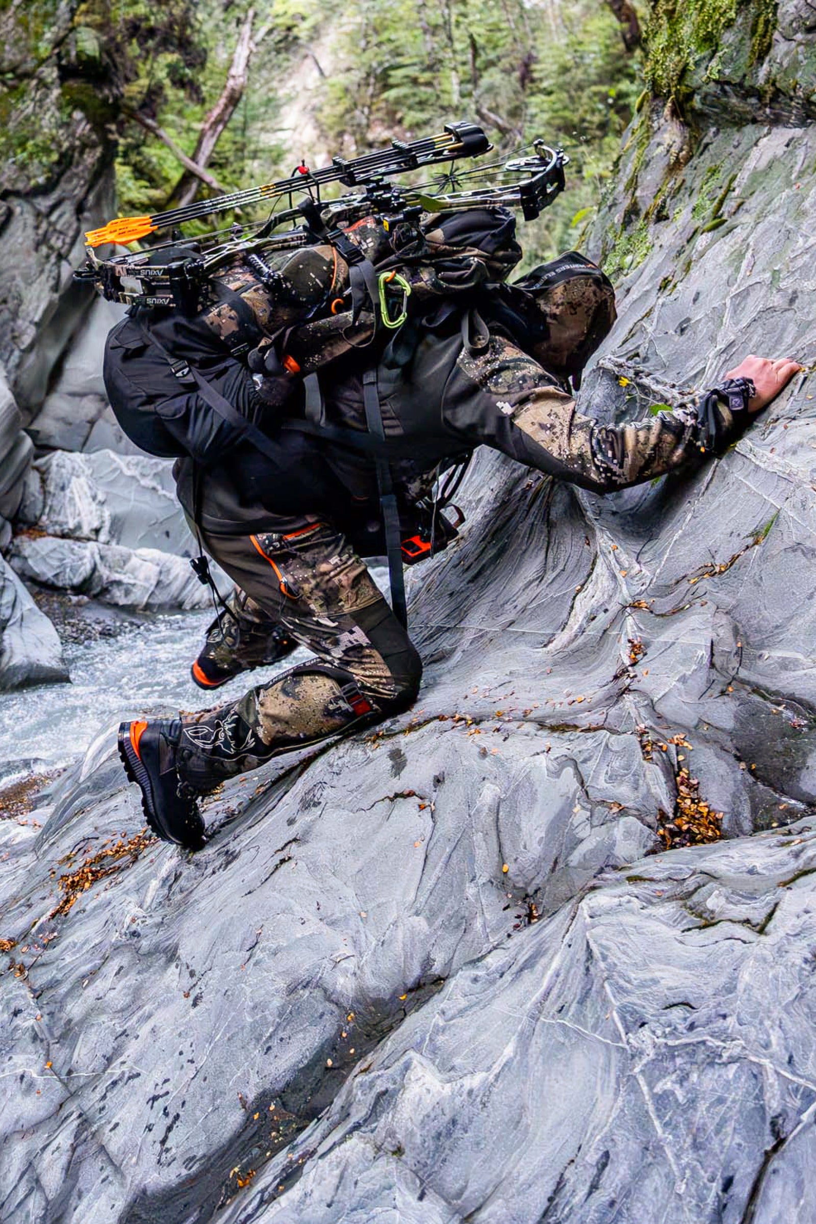 Person in camouflage gear climbing a rocky mountain with archery equipment.