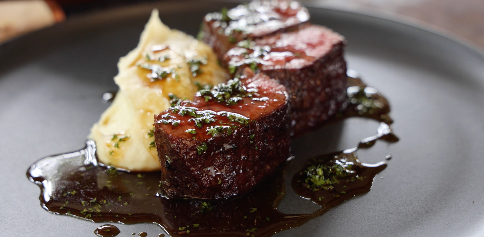 Two pieces of cooked meat with a side of mashed potatoes on a dark plate.
