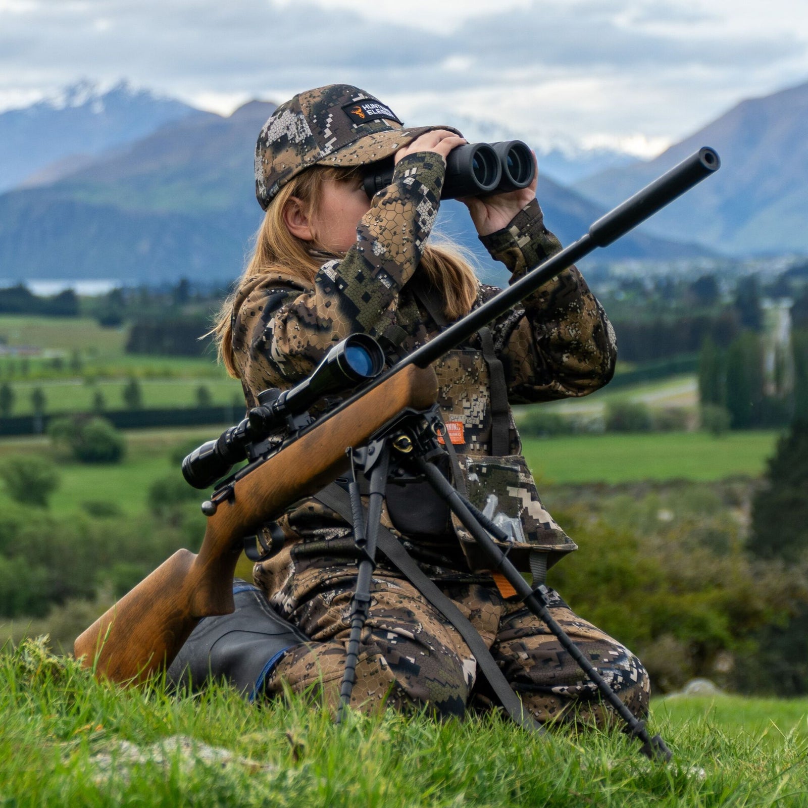Person in camouflage gear with a rifle and binoculars in a scenic outdoor setting