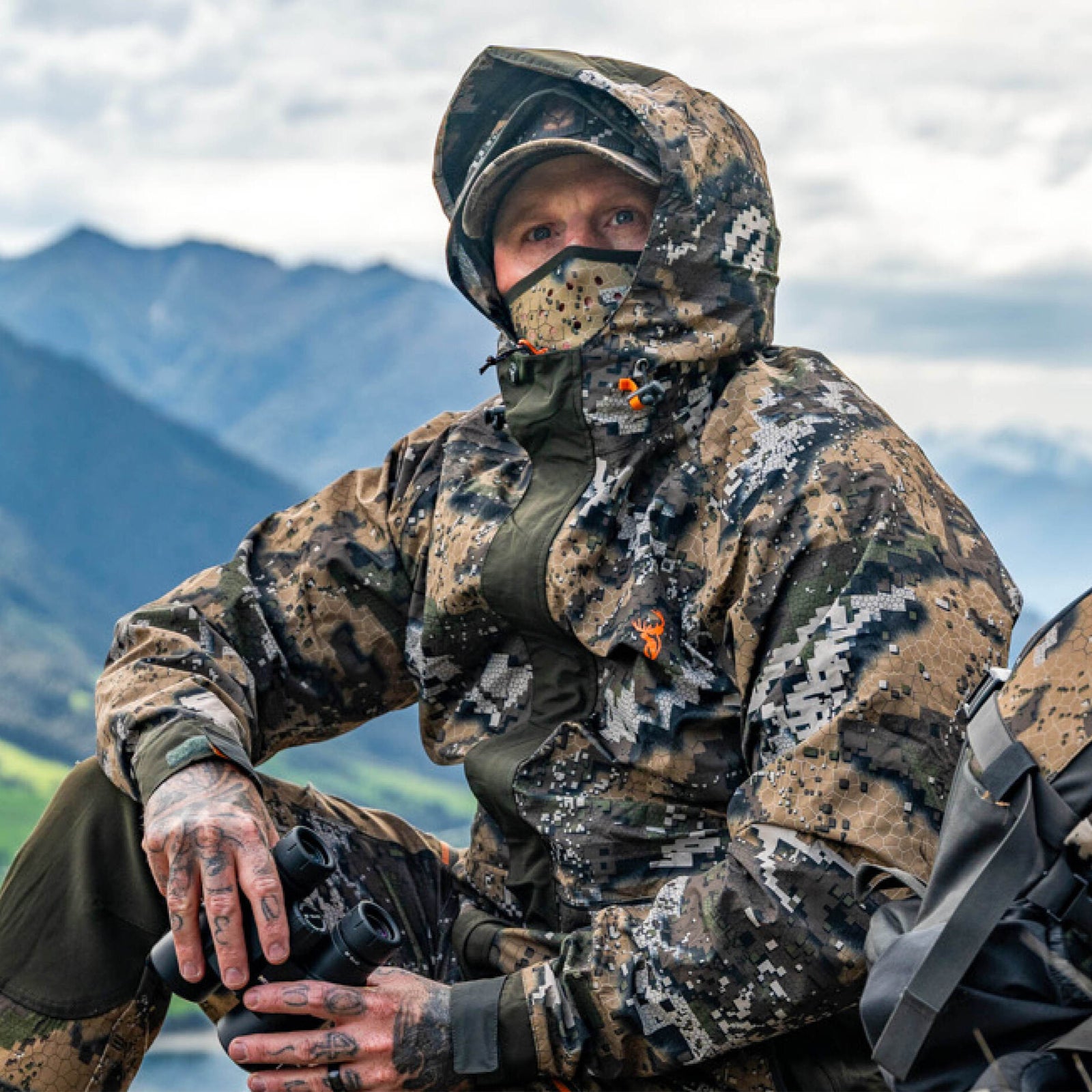 Person in camouflage hunting gear sitting outdoors with mountains in the background